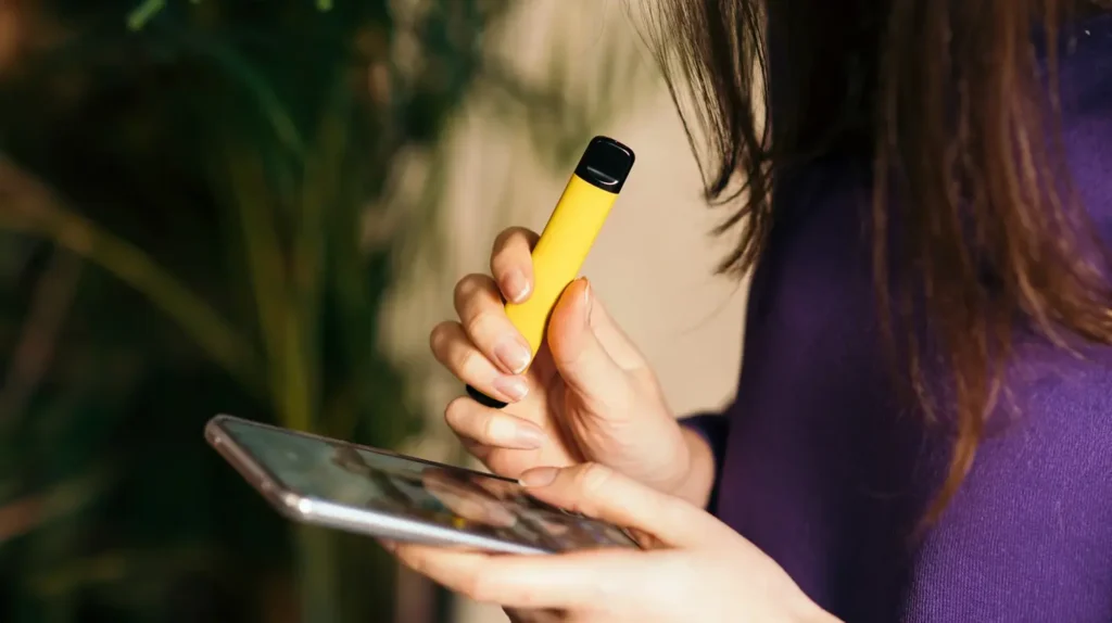 Une femme tient une cigarette électronique jetable jaune et utilise un smartphone. L'image illustre la question "Comment savoir si votre Puff est finie" en montrant un moment d'utilisation courante de la puff.