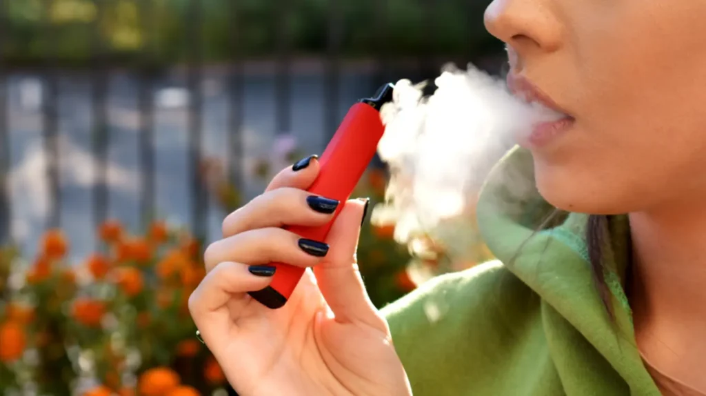 Jeune femme fumant une puff rouge, montrant comment fumer une puff. La vapeur est inhalée dans un cadre extérieur avec des fleurs en arrière-plan.