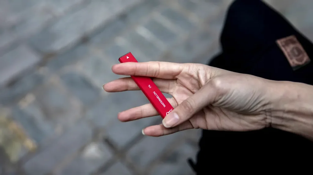 "Main tenant une puff bar rouge vif en gros plan" - Une main féminine tient une puff bar rouge avec la mention "watermelon" visible sur le dispositif, sur un fond de rue pavée. L'image met en évidence la simplicité et l'élégance de la puff bar, idéale pour une utilisation discrète et mobile.